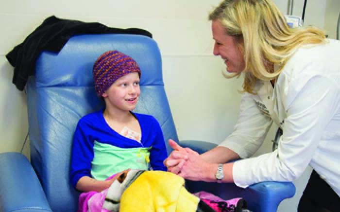 A small girl in a hospital chair with a hat on, talking to a nurse.